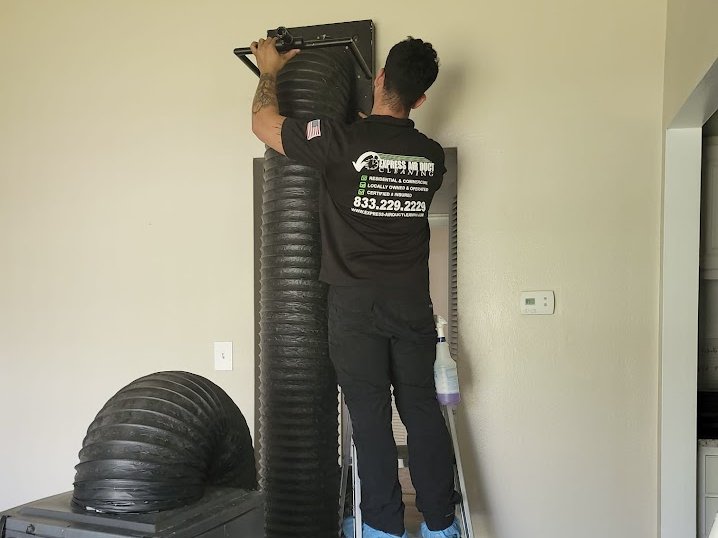 Express Air Duct Cleaning technician in a company shirt cleaning a wall vent with a duct hose